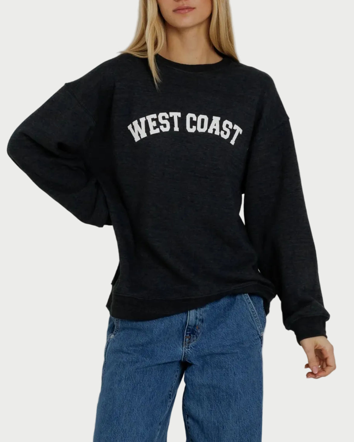 A person is wearing the Sunny Somewhere West Coast Oversized Crew by Sunny Somewhere—a black oversized sweatshirt with “WEST COAST” in white letters on the front—paired with blue jeans, standing against a plain background.