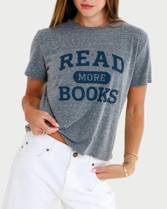A woman wears the Sunny Somewhere Read More Books Tee with a relaxed boxy fit, styled with white pants and gold bracelets, standing against a light background.