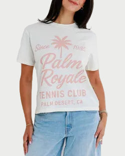 Woman wearing a white t-shirt with 'Palm Royale Tennis Club' text and blue shorts on a light gray background