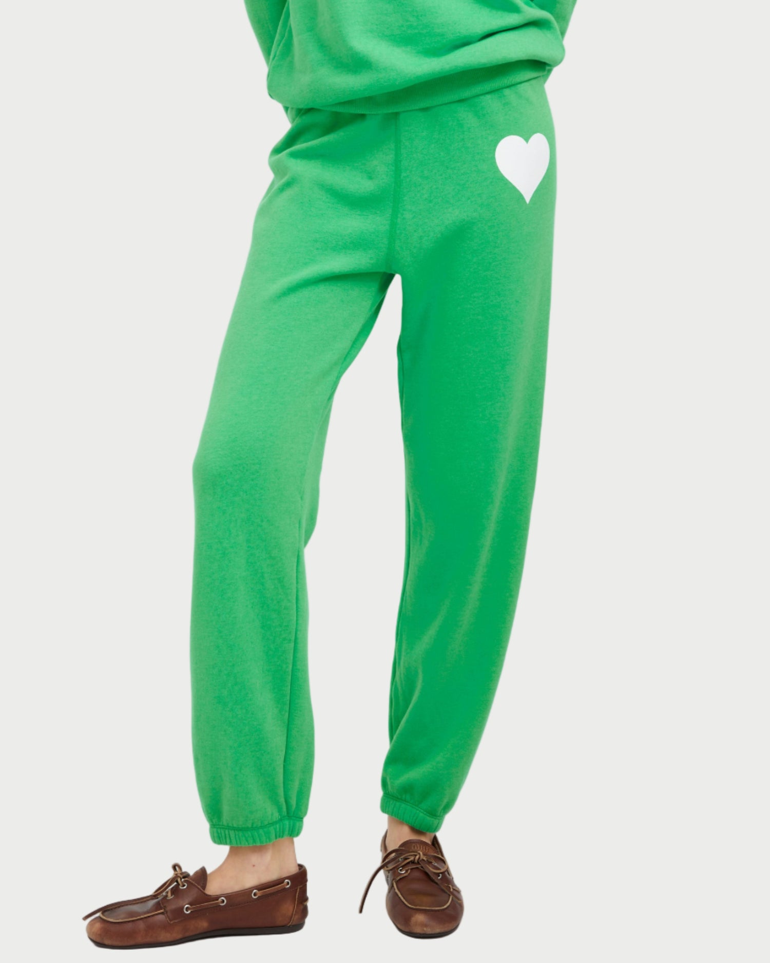 A person wears SPRWMN Heart Sweatpants in bright green with a white heart graphic on the upper left thigh, paired with brown moccasin-style shoes. The SPRWMN joggers pop against the plain white background.