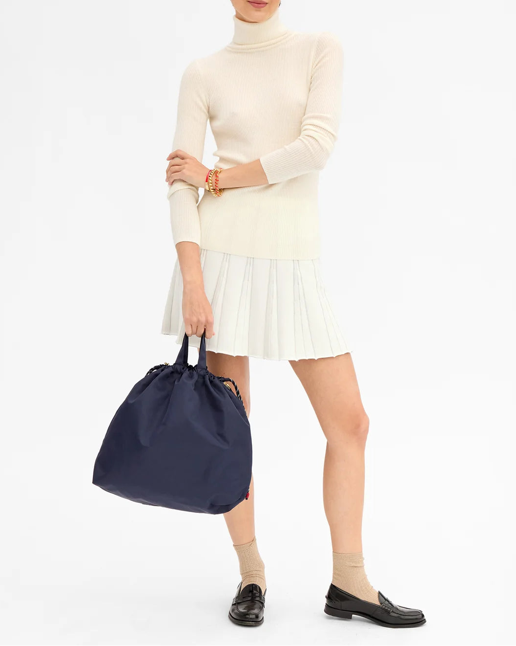 A person wearing a cream turtleneck sweater and pleated skirt holds the Clare V. Nylon Sac Tout by CLARE V., a large navy recycled polyester bag. They wear tan socks with black loafers. Their head is not visible.