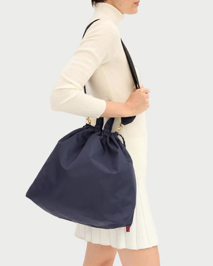 A person wearing a white long-sleeve top and skirt carries the Clare V. Nylon Sac Tout by CLARE V.—a large navy blue vegan carryall featuring black straps and an adjustable crossbody strap—against a plain light background.