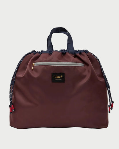 A dark burgundy drawstring bag with navy blue handles and braided side straps, featuring a front zip pocket and a black Clare V London brand patch.