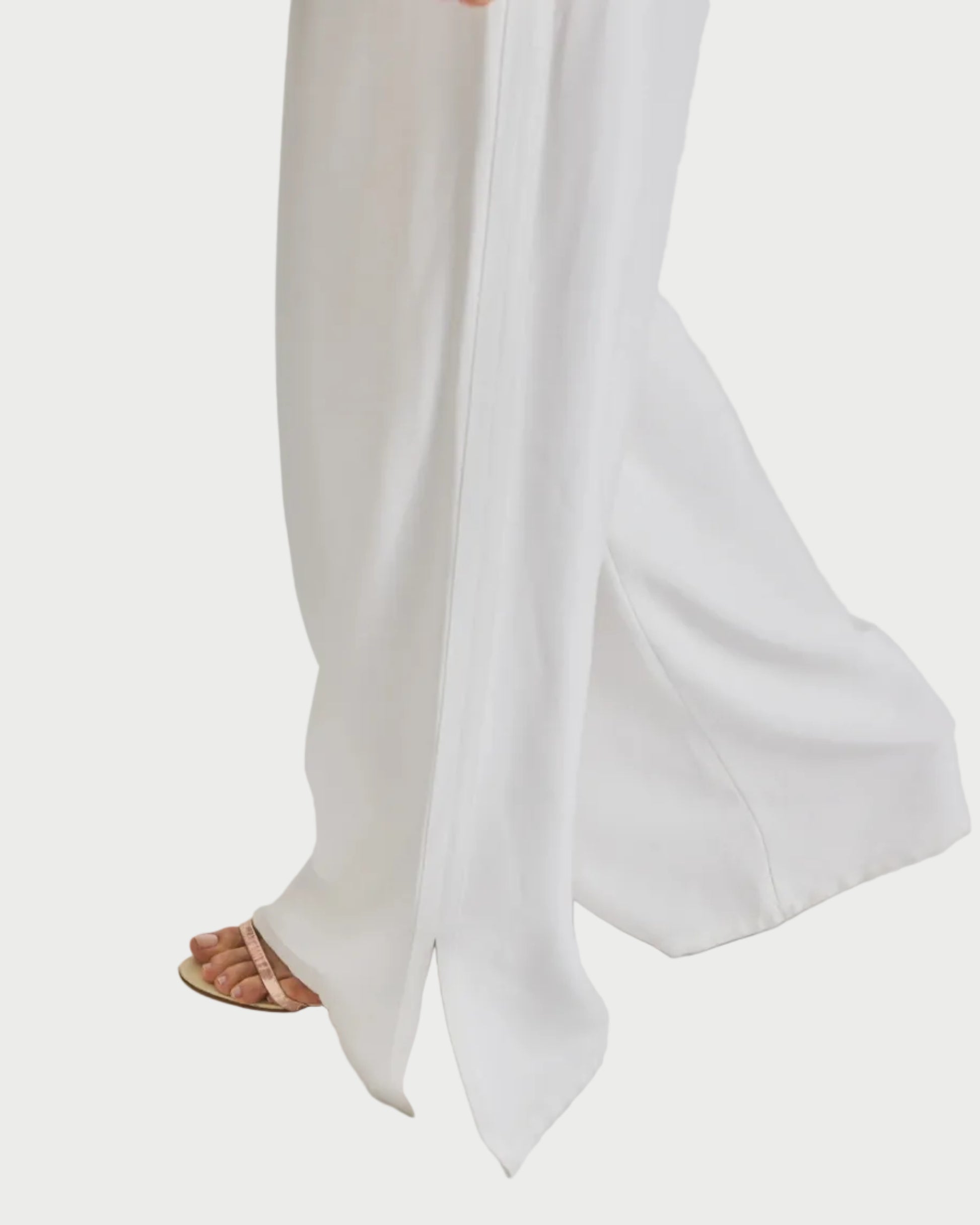Wearing the Saint Art NY Neve Tuxedo Pant with a wide-leg silhouette and slit hem, paired with beige open-toe sandals, a model walks against a plain white background.