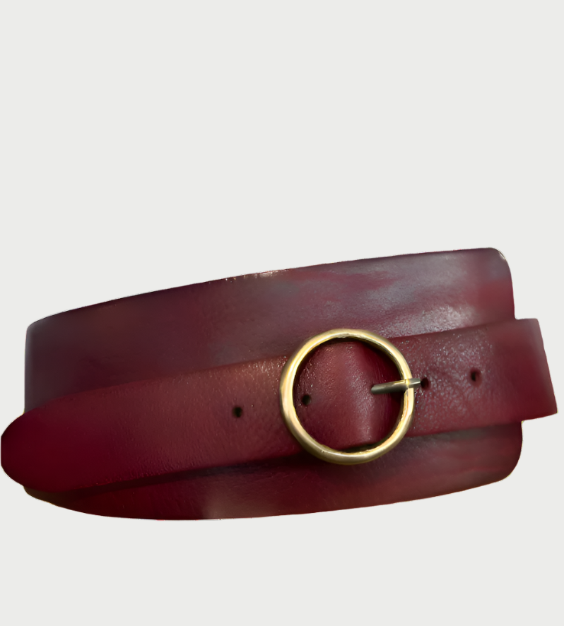The Wide Contour Belt by Motif56, a dark red leather accessory with a simple gold round buckle, rests coiled on a plain background. Its smooth texture and high-quality craftsmanship evoke 70's boho-chic vibes.