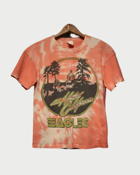 The Madeworn Eagles Tee is a vintage-fit, 100% cotton tie-dye shirt in orange and cream, featuring "Hotel California" and "Eagles" text alongside palm trees and a silhouetted building for a retro look.