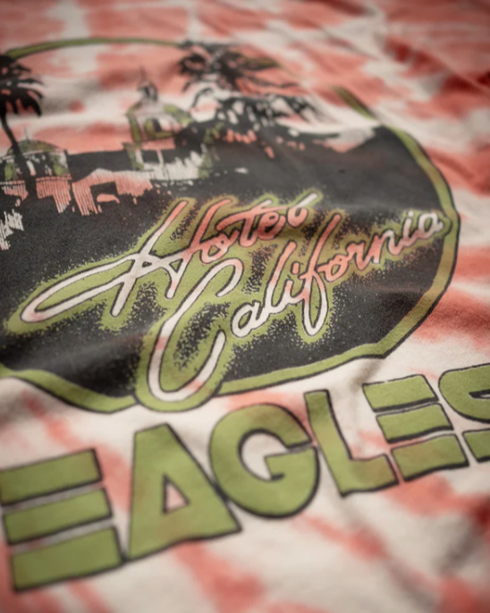 A close-up of Madeworn's "The Eagles Tee" reveals its vintage fit and design, with "Hotel California Eagles" in stylized fonts. It features palm trees and a mission-style building on 100% cotton, offering a retro California vibe.