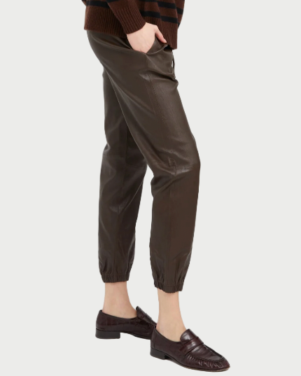 A person wearing SPRWMN Leather Drawstring Jogger pants by SPRWMN, paired with a brown striped sweater and brown loafers, stands with one hand in their pocket against a white background.