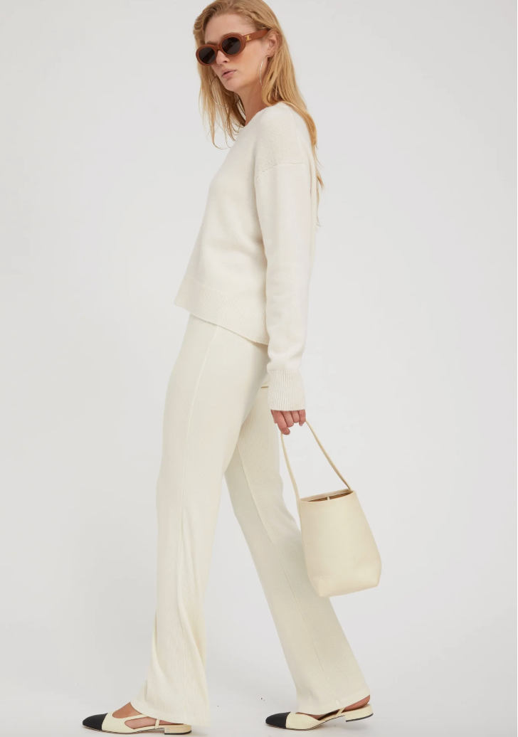 A woman in profile wears the SPRWMN Boxy Cashmere Sweater in Chalk, matching wide-leg pants, sunglasses, and two-tone flats, holding a light beige handbag against a plain white background.