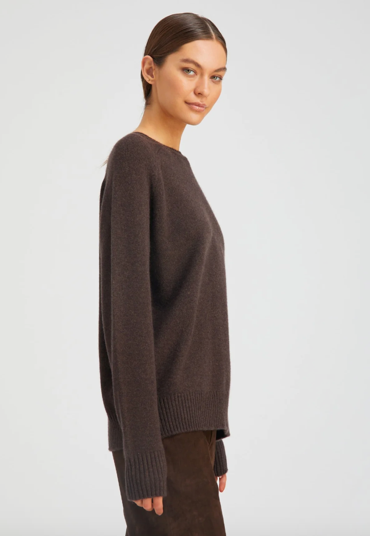 A woman with straight brown hair tied back wears the SPRWMN Cashmere Boyfriend Sweater in dark brown, paired with a matching skirt. She stands sideways and looks at the camera against a plain light background.