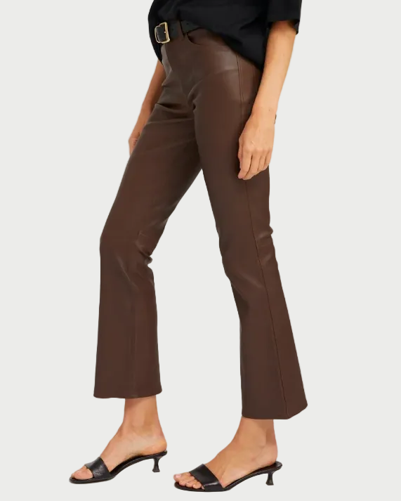 A person wearing SPRWMN 5 Pocket Ankle Flare pants in brown French lambskin leather, paired with a black top, black belt with a gold buckle, and black heeled sandals, stands against a plain white background.