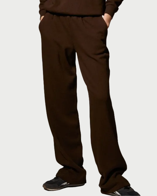 A woman modeling a matching dark brown lounge set consisting of a half-zip pullover sweatshirt and wide-leg sweatpants, styled with dark sunglasses and sneakers against a neutral gray studio background.