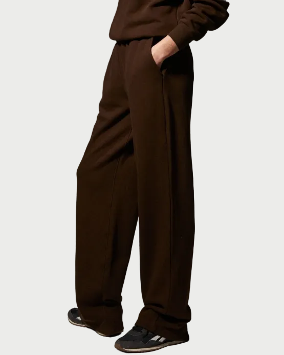 A person wearing SPRWMN Straight Leg Sweatpants in brown cotton-fleece with an adjustable drawstring waist, paired with a matching brown sweatshirt and black sneakers featuring white stripes, stands with one hand in their pocket against a plain background.
