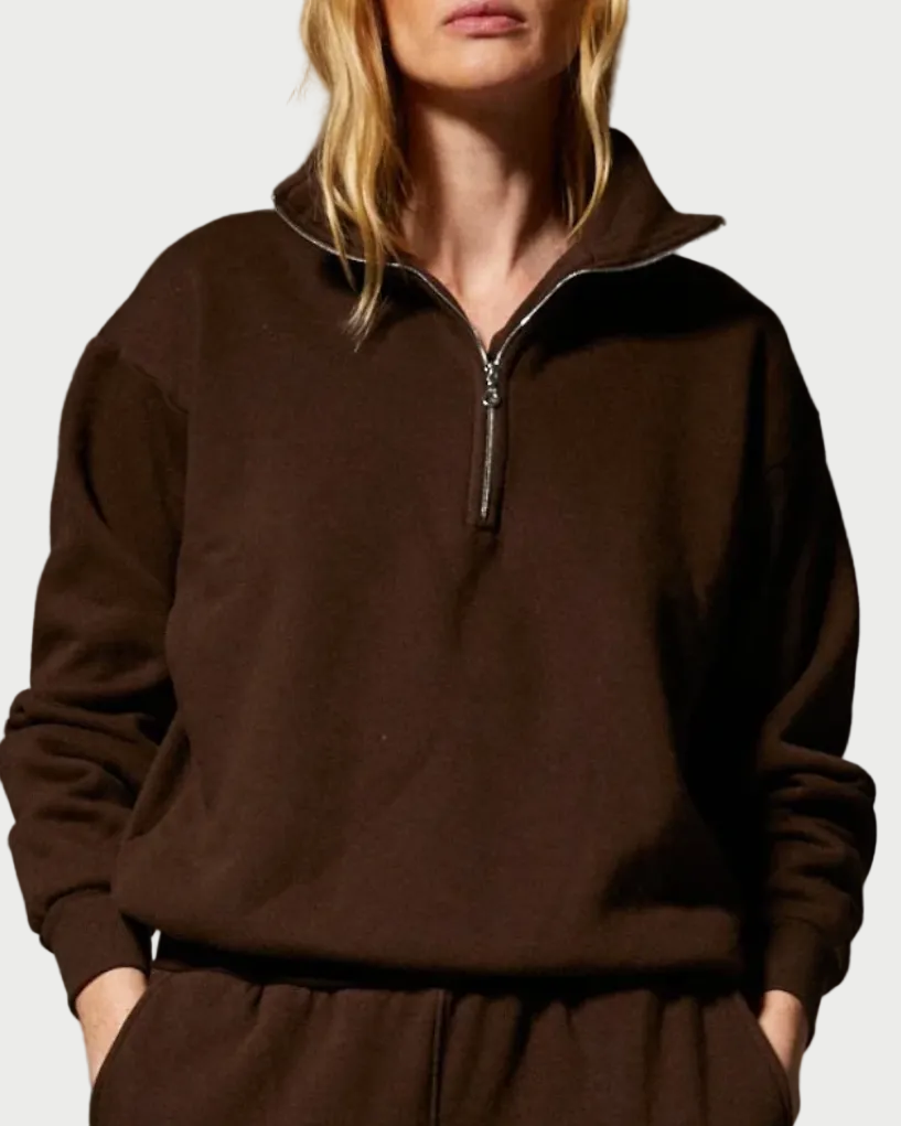 A woman with blonde hair and her hands in her pockets stands against a plain light background, wearing a dark brown SPRWMN 1/4 Zip Sweatshirt in a relaxed fit with matching sweatpants.