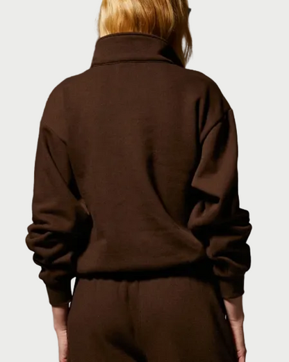 A person with long blond hair is shown from the back, wearing the SPRWMN 1/4 Zip Sweatshirt—a dark brown, relaxed fit cotton-fleece top with a high collar by SPRWMN—and matching pants, standing against a plain light background.