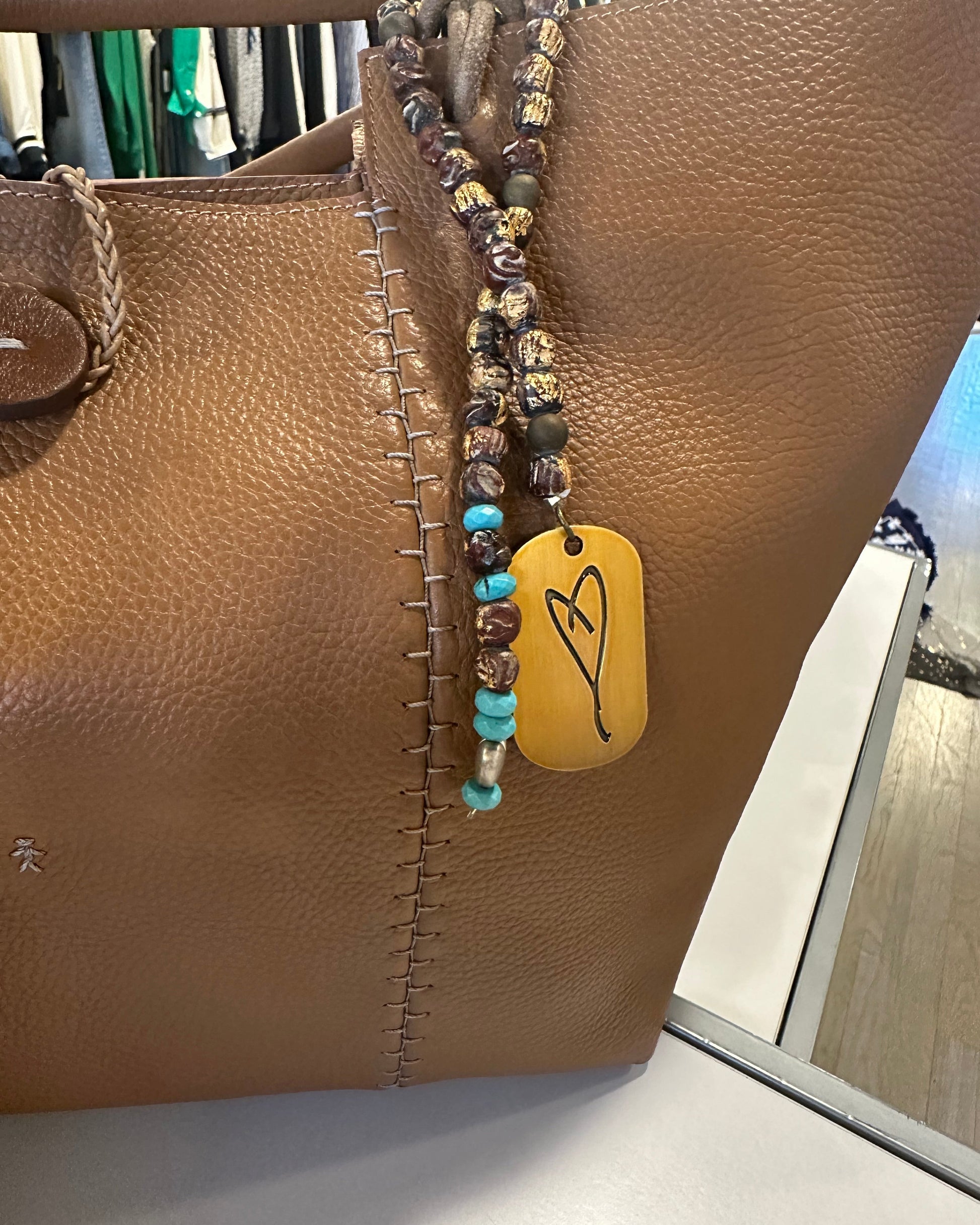 On display is a MustHave accessory: Simply Eartha's Here's To Life Bag Charm. It's paired with an antique gold beaded necklace featuring a heart pendant, enhancing its charm. In the softly blurred background, clothing subtly complements this elegant setting.