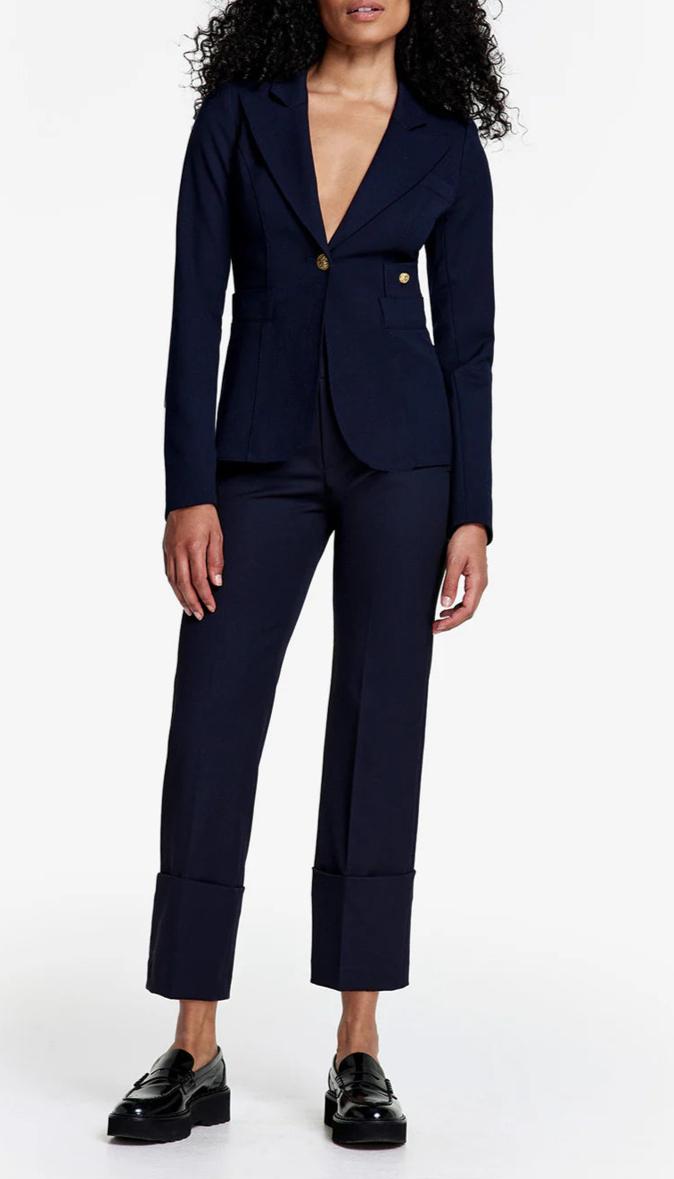 A person stands against a plain white background wearing the Smythe Full Back Duchess Blazer in Navy, styled with tailored cropped trousers and black loafers.