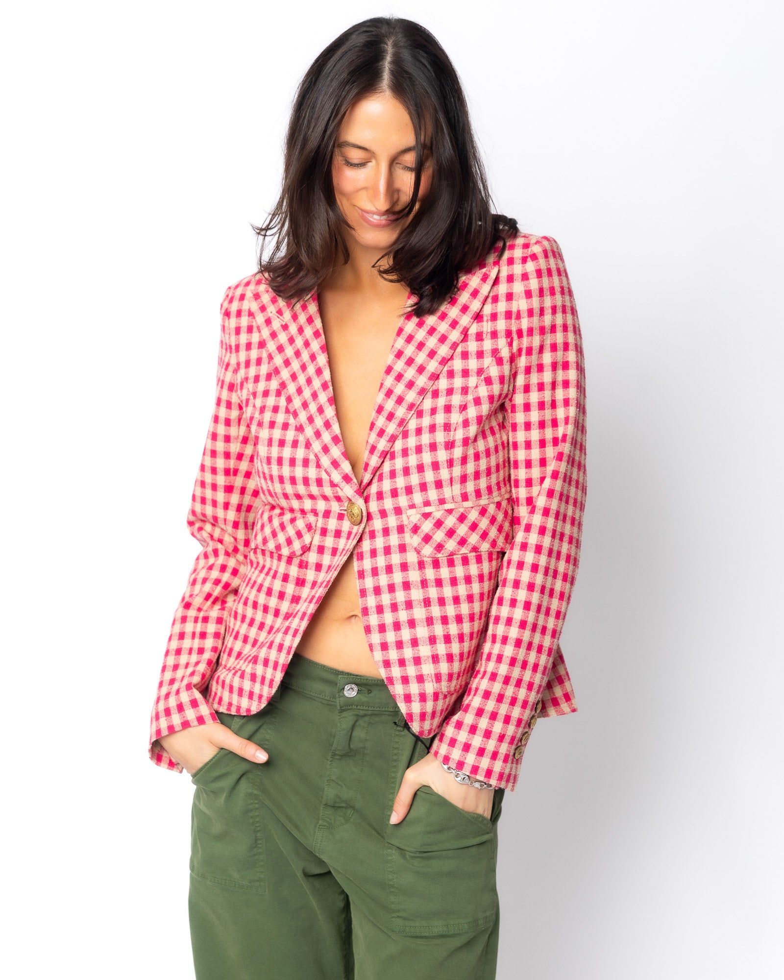 A woman with dark hair smiles and looks down, wearing the Smythe Patch Pocket One Button Blazer by Smythe with no shirt underneath and green pants, standing against a white background with her hands in her pockets.