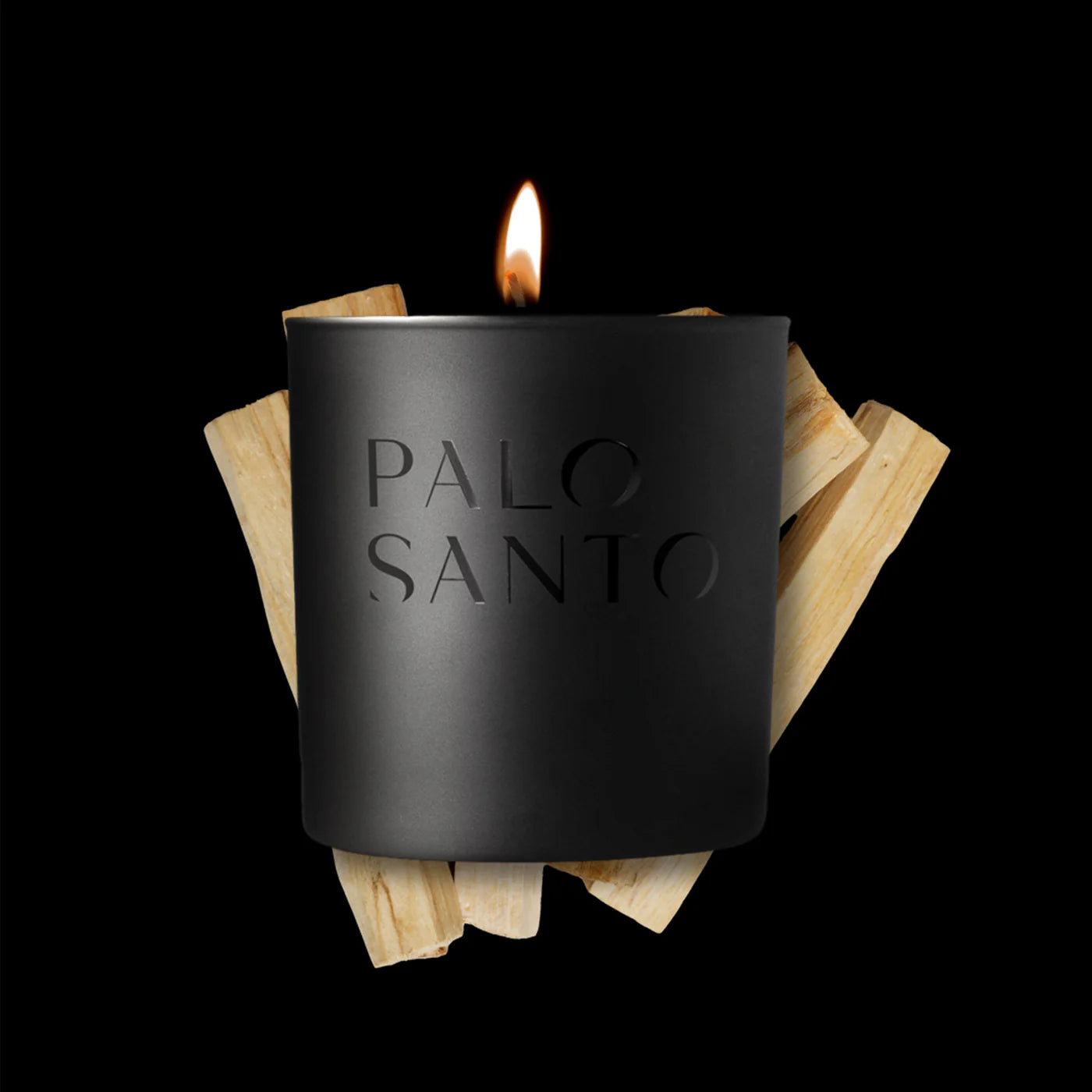 The Studio Stockhome Palo Santo Candle, a black luxury soy candle with a glowing flame, sits before light palo santo wood sticks on a black background—ideal for fans of natural candles by Studio Stockhome.