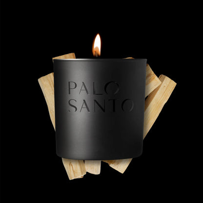The Studio Stockhome Palo Santo Candle, a black luxury soy candle with a glowing flame, sits before light palo santo wood sticks on a black background—ideal for fans of natural candles by Studio Stockhome.