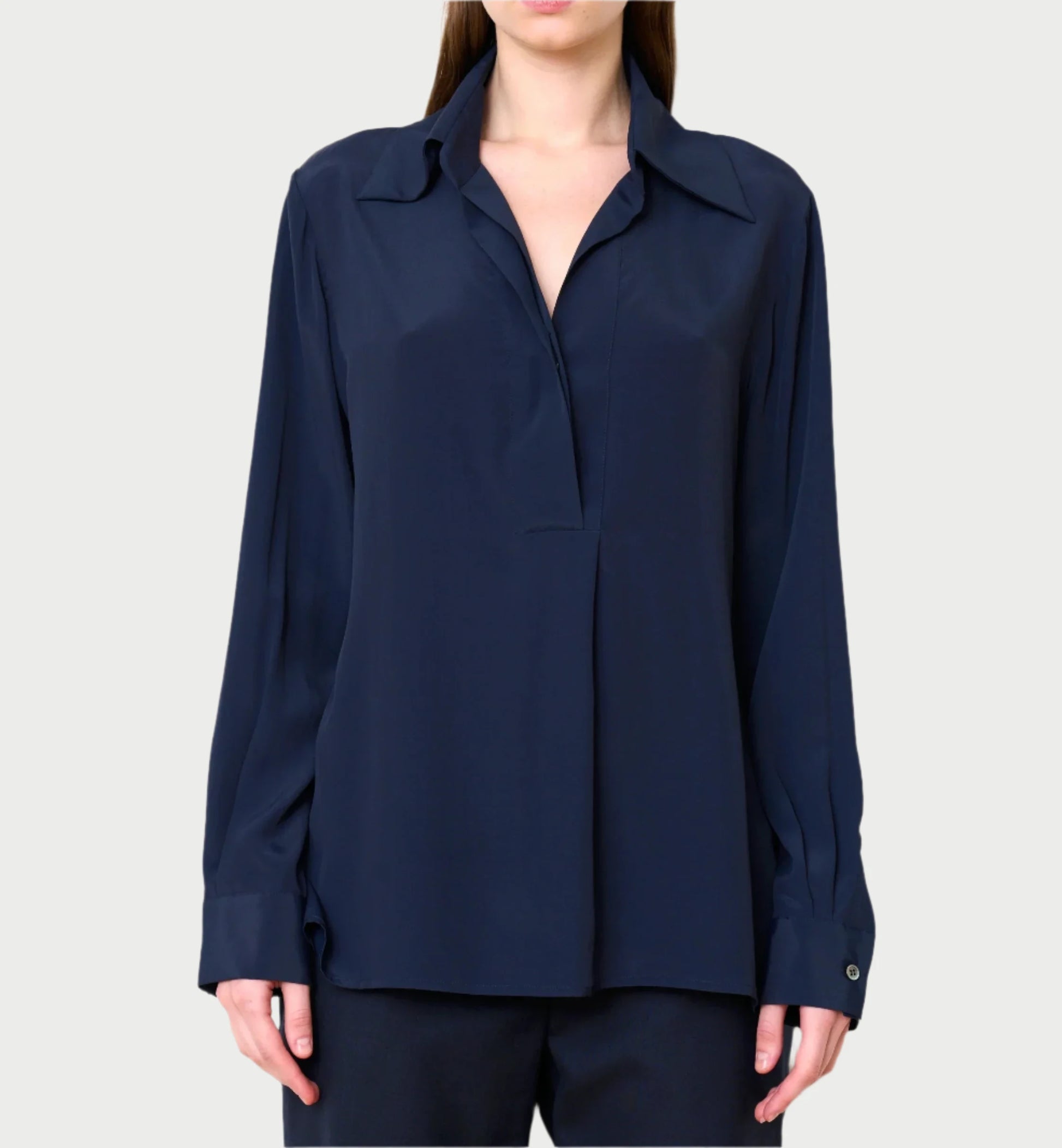 A person wears the Standard Blouse by WE-AR4, a dark blue, long-sleeved collared blouse made from luxury deadstock materials. It has a loose fit and a subtle open V-neck design, featuring buttoned cuffs. The background is a neutral light gray.