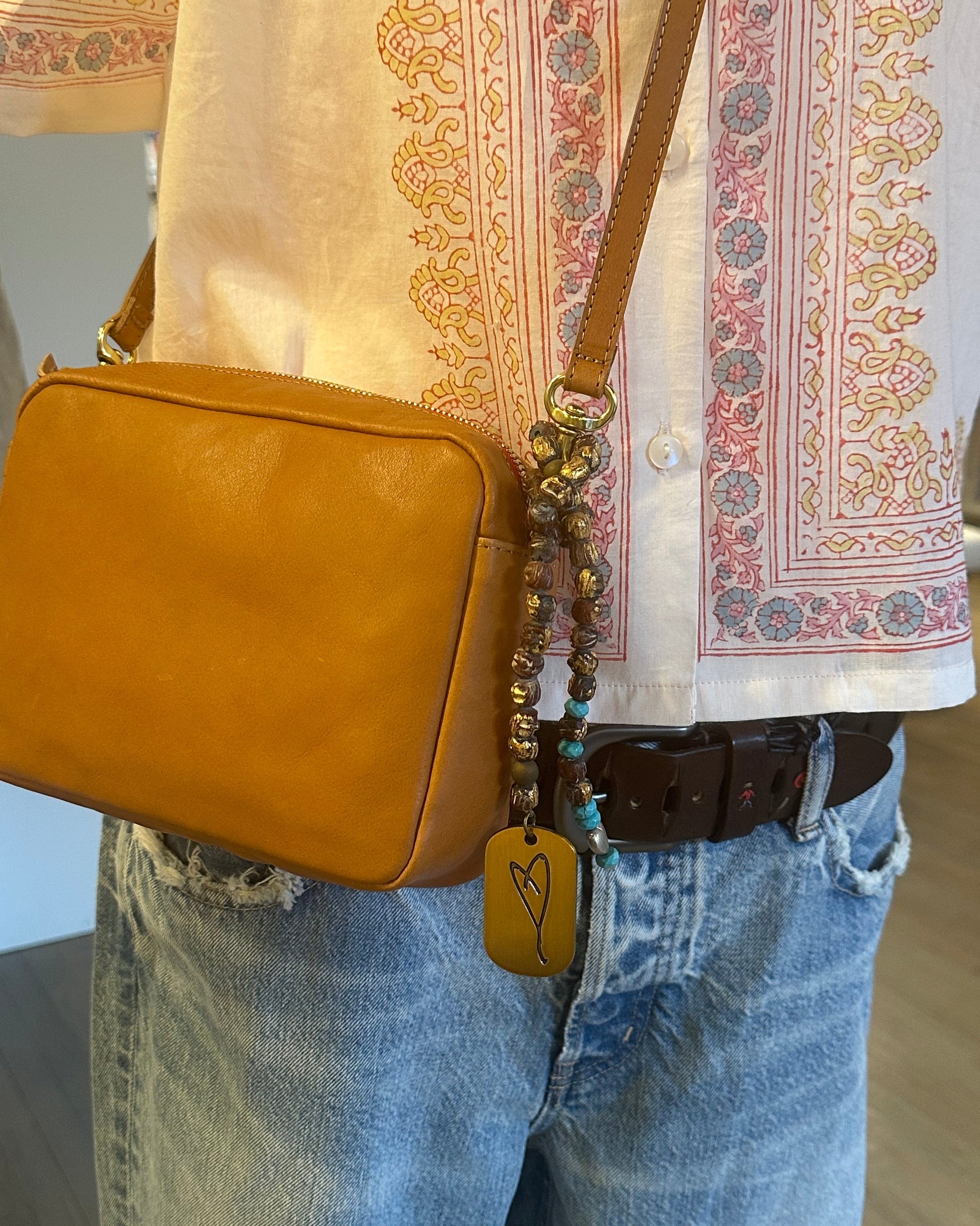 A person in a patterned shirt, blue jeans, and a brown belt carries Simply Eartha's Here's To Life Bag Charm, featuring an antique gold strap with beads and a heart-engraved accent.