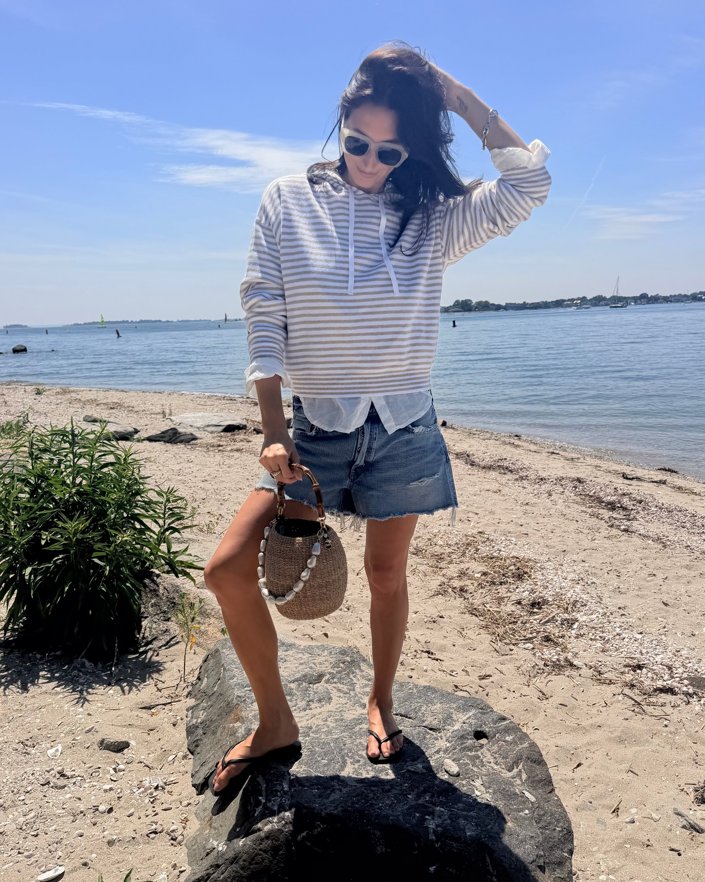 A woman in sunglasses and denim shorts stands on a rock at the beach, wearing the One Grey Day Striped Warwick Hoodie from One Grey Day, paired with flip-flops and holding a straw bag, with water and blue sky in the background.