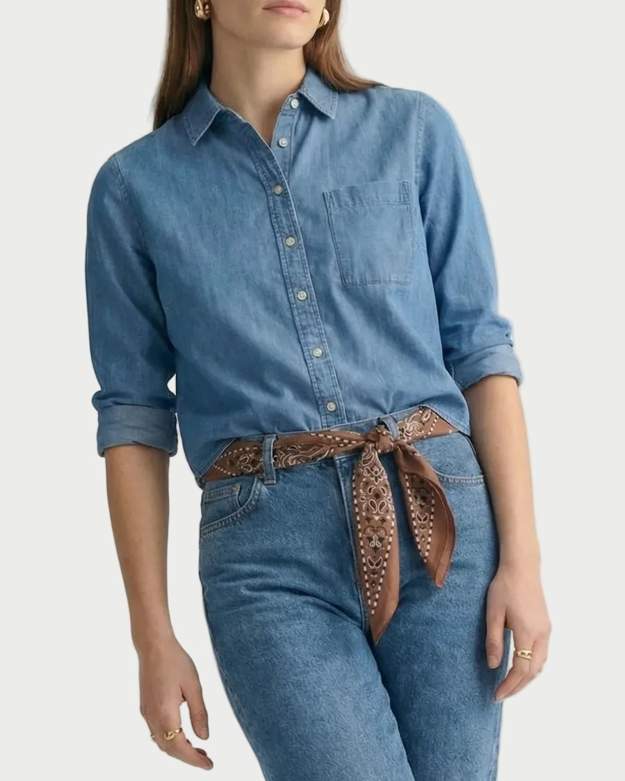 A person wears The Shirt Long Sleeve Boxy Crop, a cotton, button-down denim shirt tucked into blue jeans with a brown patterned scarf tied as a belt.