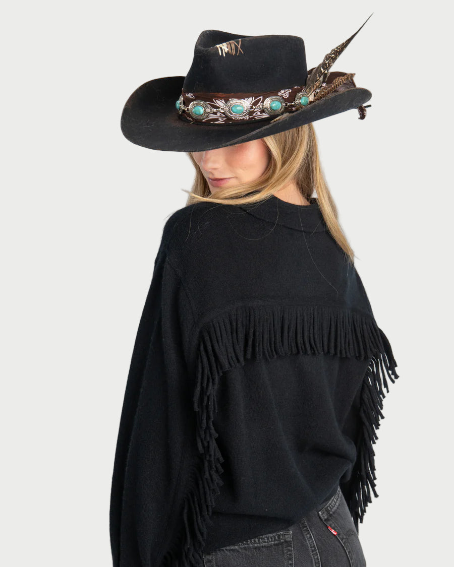 Person wearing a black cowboy hat with feather and turquoise stones, and a black fringe sweater on a gray background.