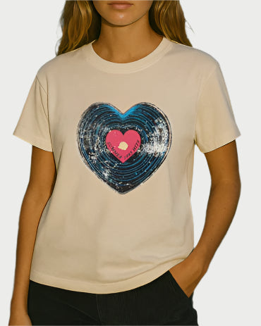 A woman wears the Sunny Somewhere Record Everyday Tee, a beige shirt with a large blue heart-shaped record graphic, featuring a pink heart and "SOCIAL IMPACT" in white. Perfect with vintage denim.