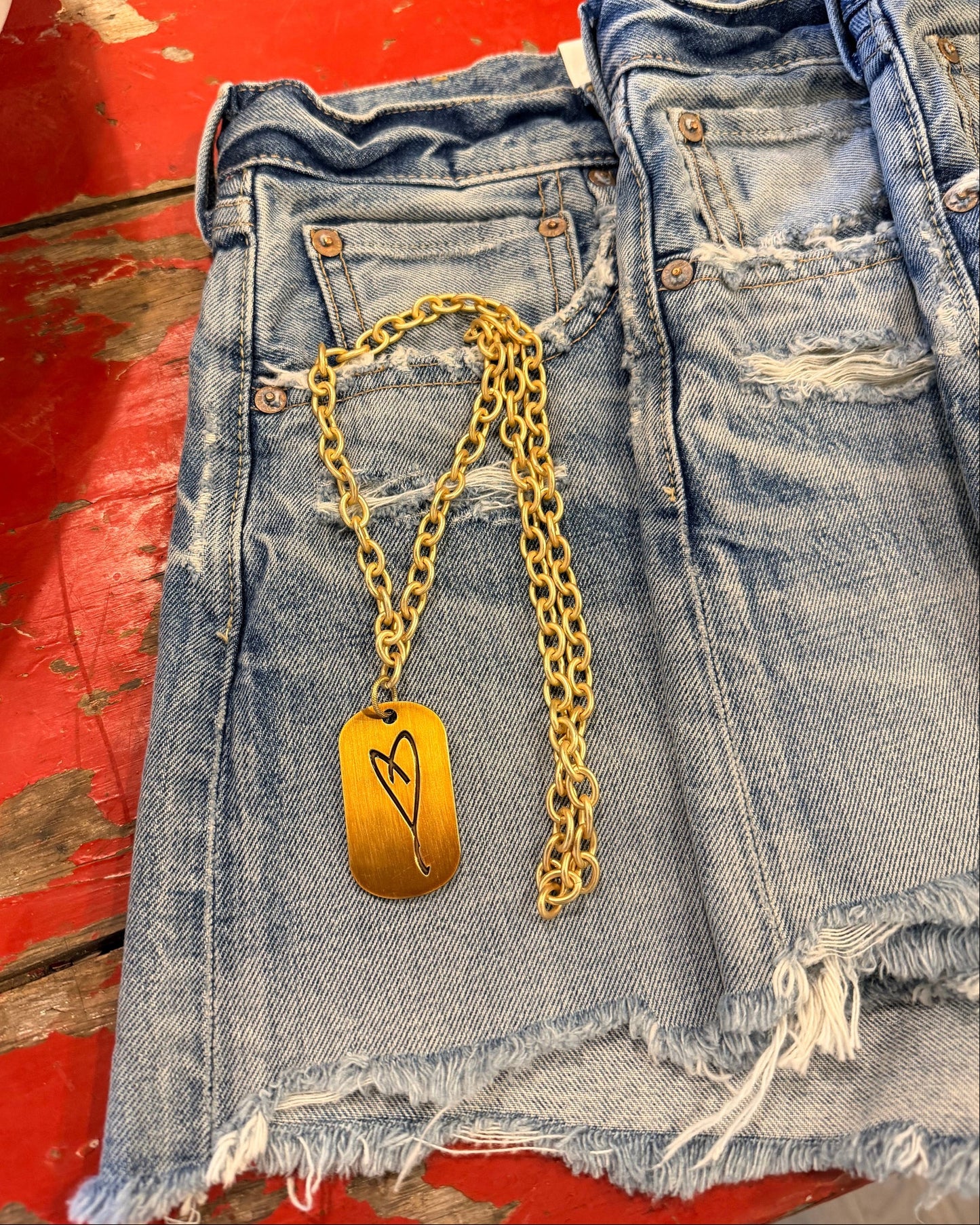 The Simply Eartha "Here's To Life/ EK Heart" gold necklace with a rectangular heart pendant lies on frayed blue denim shorts on a distressed red wooden surface.