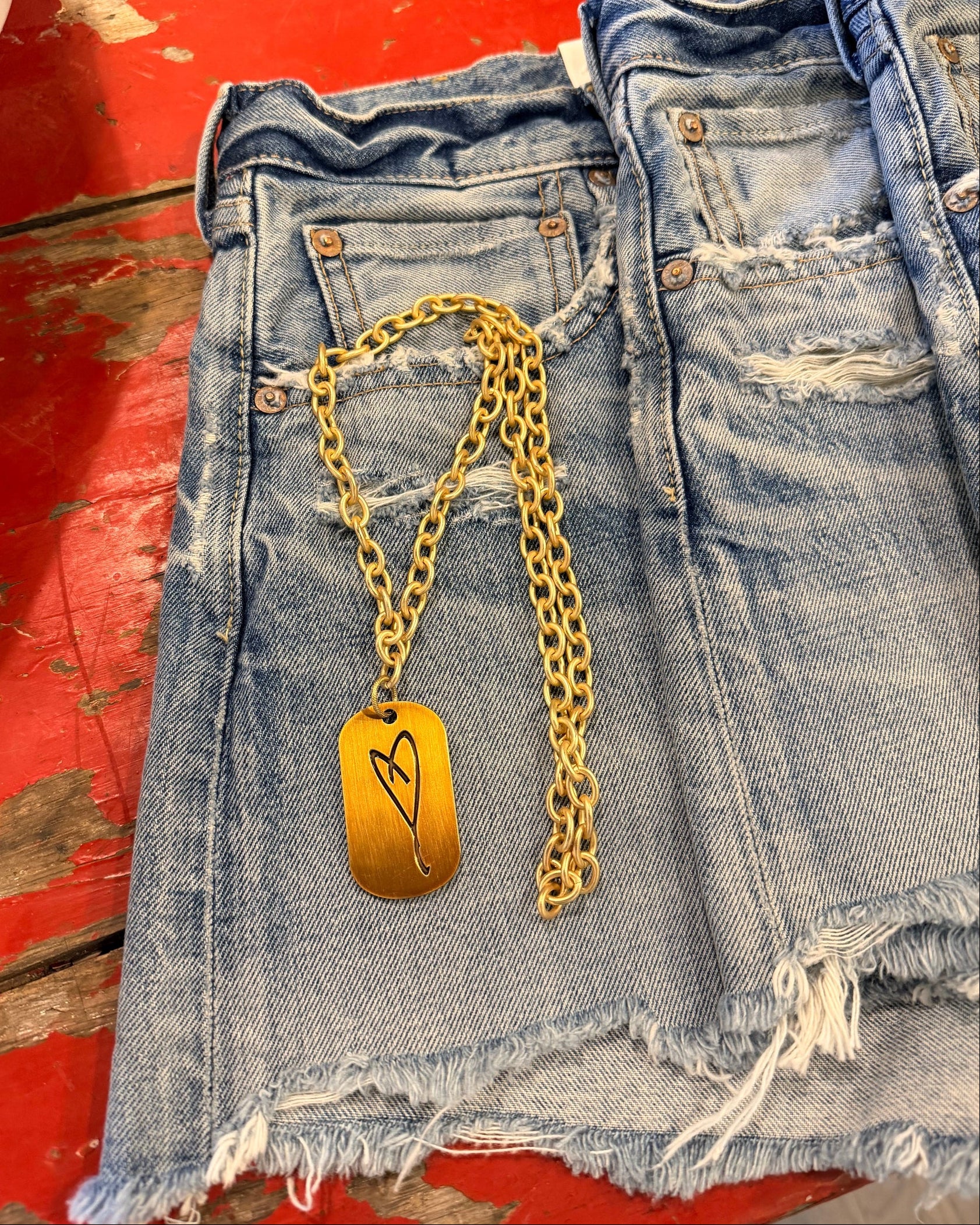 The Simply Eartha "Here's To Life/ EK Heart" gold necklace with a rectangular heart pendant lies on frayed blue denim shorts on a distressed red wooden surface.