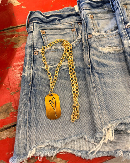 The Simply Eartha "Here's To Life/ EK Heart" gold necklace with a rectangular heart pendant lies on frayed blue denim shorts on a distressed red wooden surface.