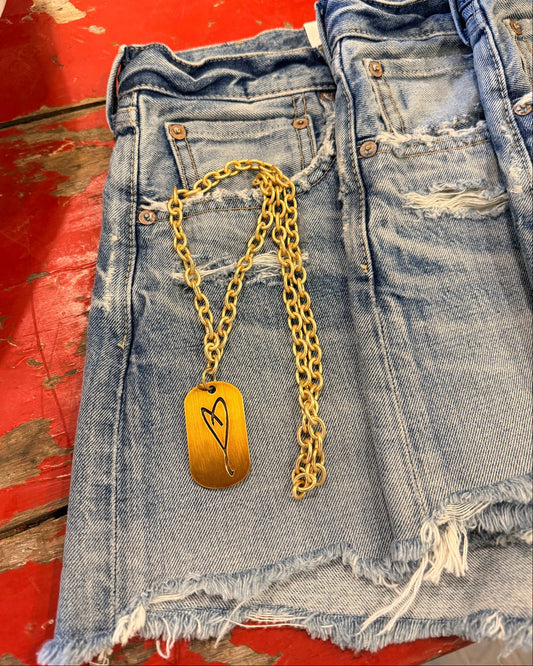 The Simply Eartha "Here's To Life/ EK Heart" gold necklace with a rectangular heart pendant lies on frayed blue denim shorts on a distressed red wooden surface.