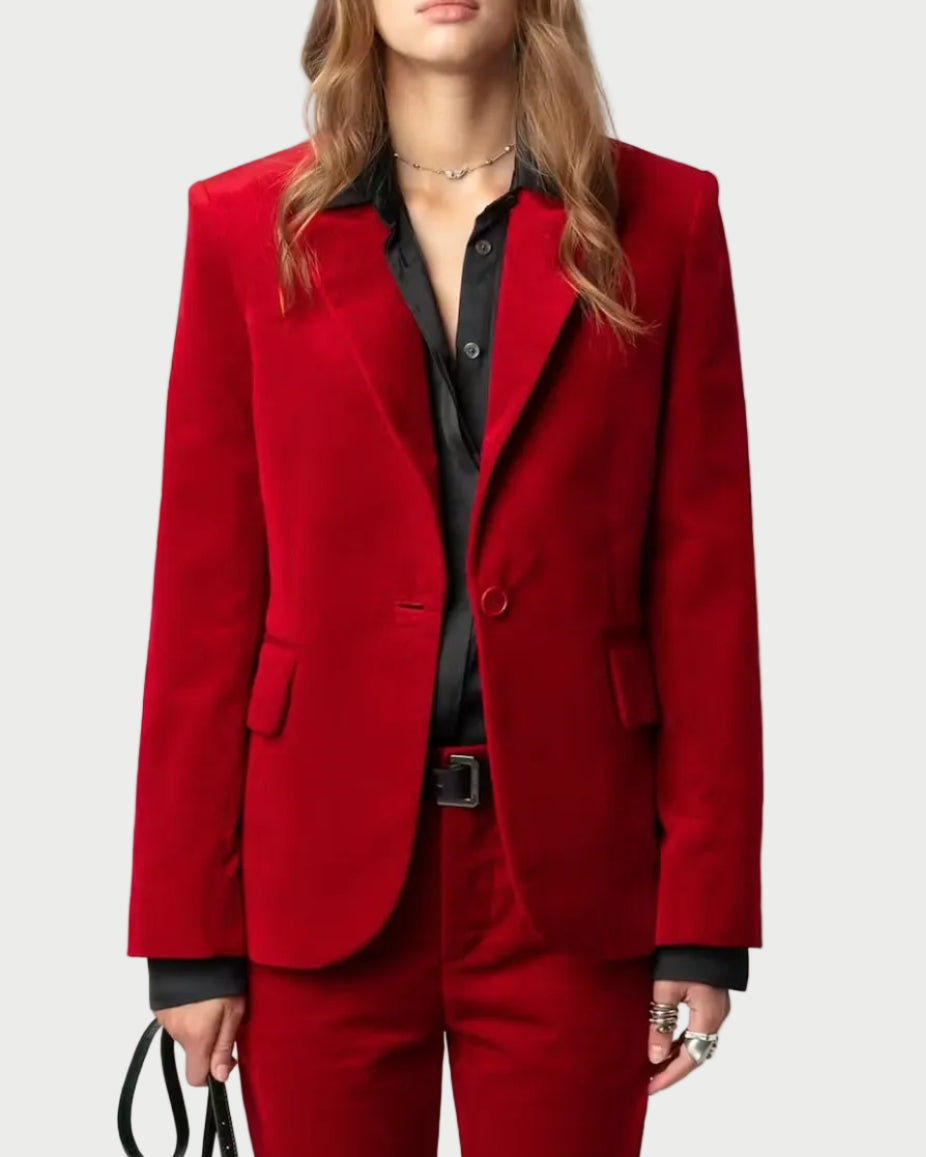 A model wearing a structured red velvet pantsuit with a matching blazer and trousers, styled with a black button-down shirt, black sunglasses, and holding a black handbag against a light gray background.