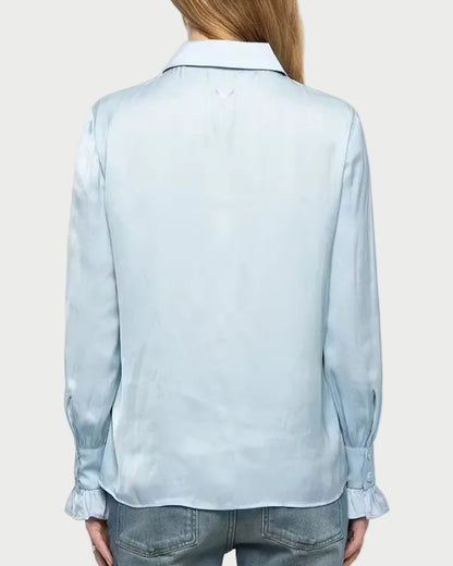A woman viewed from behind wearing a light blue long-sleeve button-up shirt and blue jeans against a plain light gray studio background.
