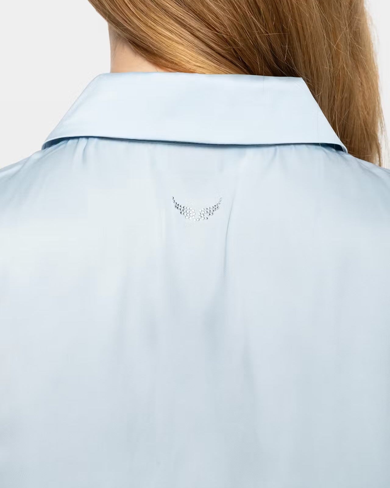 Close-up back view of a light blue collared shirt worn by a model, showing a small silver rhinestone wing emblem centered below the collar.