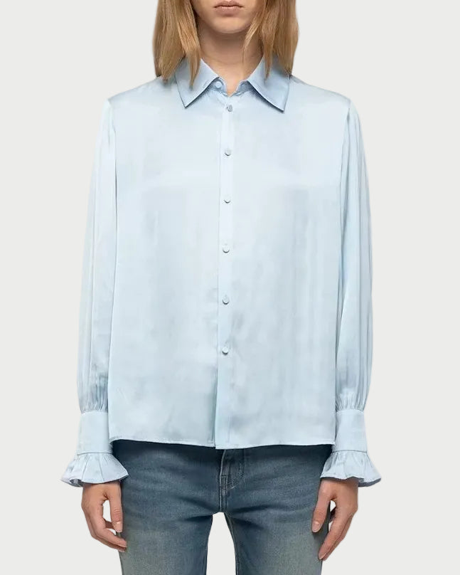 A model stands against a light gray studio background wearing a light blue long-sleeve button-up shirt, blue jeans, and white sneakers.