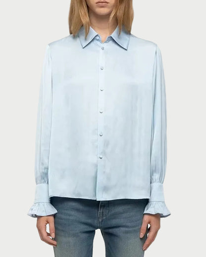 A model stands against a light gray studio background wearing a light blue long-sleeve button-up shirt, blue jeans, and white sneakers.