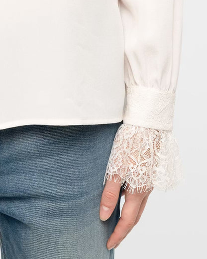 Close-up of a person wearing a cream-colored blouse with a lace ruffled cuff and blue jeans, showing the hand and part of the torso.