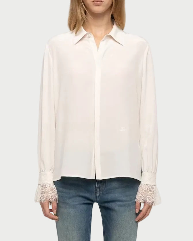 A model stands against a light gray studio background wearing a long-sleeve white button-up shirt with lace trim at the cuffs, blue jeans, and white sneakers.