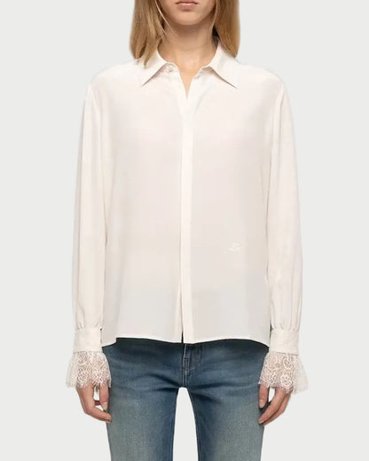 A model stands against a light gray studio background wearing a long-sleeve white button-up shirt with lace trim at the cuffs, blue jeans, and white sneakers.