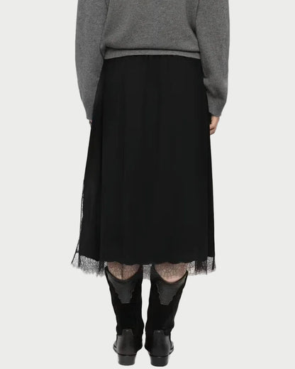 A woman viewed from behind wearing a gray knit sweater, a black midi skirt with sheer tulle hem, sheer black tights, and black ankle boots, against a light gray studio background.