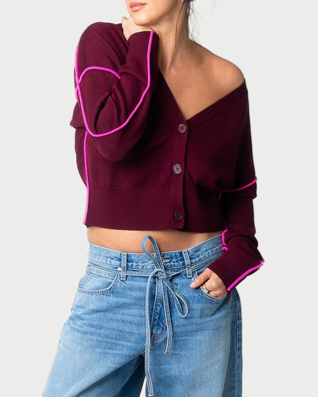 A woman wears the Ileo Cardigan by Essentiel Antwerp—a cropped wool-blend cardigan with pink trim—styled with high-waisted blue jeans, a tied belt, one hand in her pocket and the other touching her face.