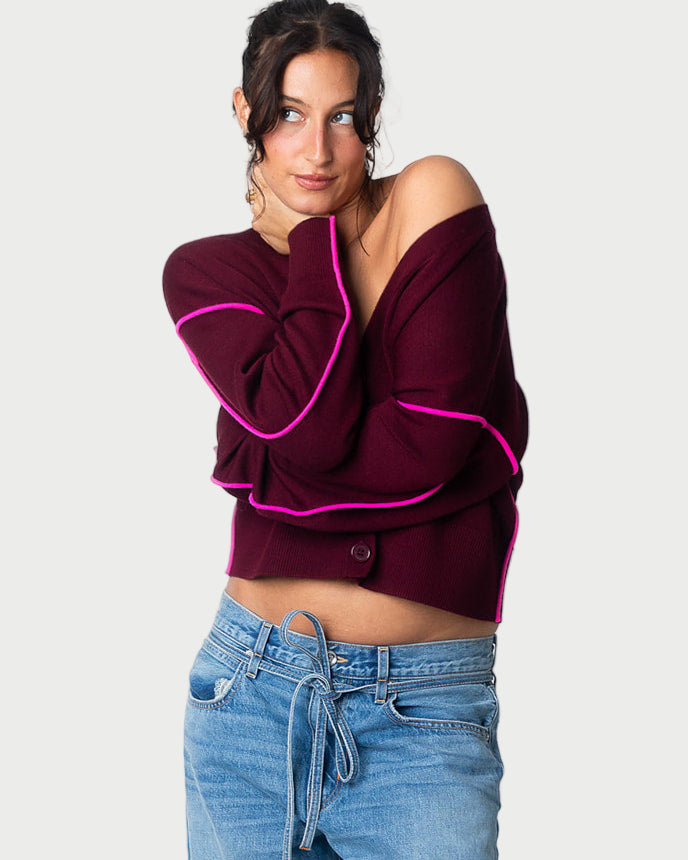 A woman with dark hair in a loose updo wears the Essentiel Antwerp Ileo Cardigan—an off-the-shoulder wool blend with pink trim—with light blue jeans. She stands against a light gray background, looking to the side with a slight smile.