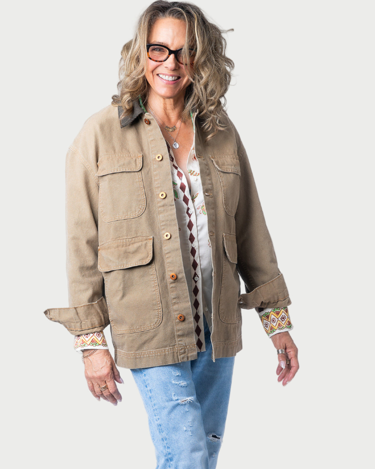 A smiling woman with wavy hair and glasses wears the SLVRLAKE Hayden Chore Jacket by SLVRLAKE with a corduroy collar over a patterned shirt and ripped blue jeans, standing against a plain white background.