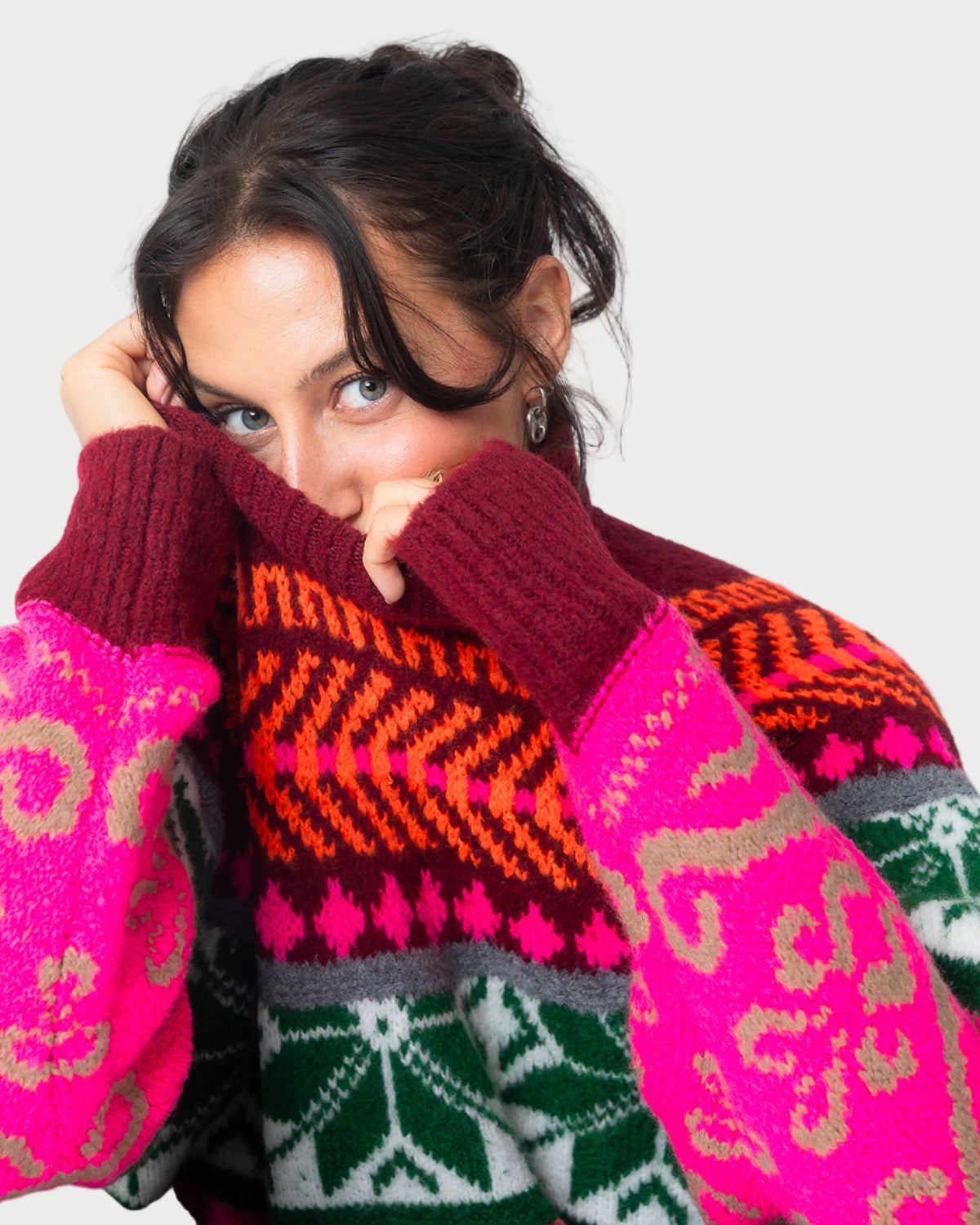 A woman with dark hair playfully peeks out from an Essentiel Antwerp Jacquard Sweater, showcasing pink, red, orange, green, and beige mélange knit patterns against a plain background.