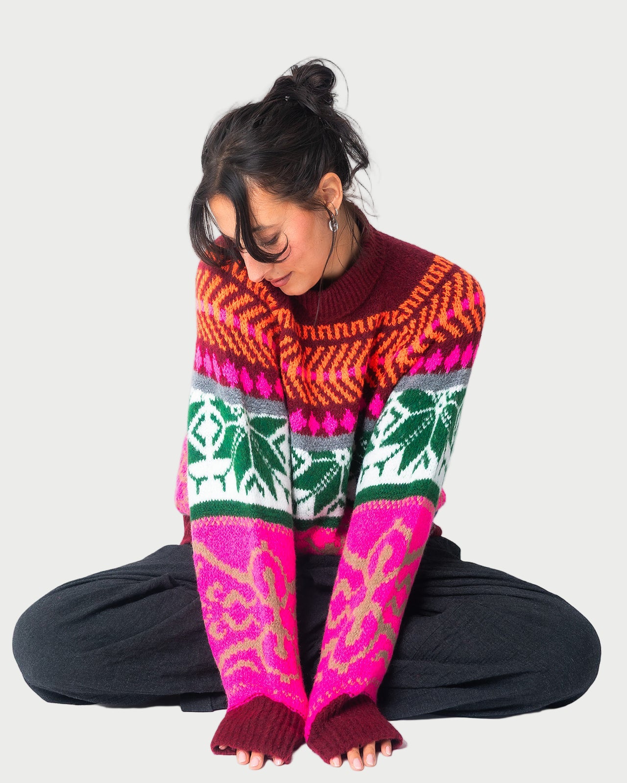 A person with dark hair in a messy bun sits cross-legged, smiling and looking down. They wear the Essentiel Antwerp Jacquard Sweater with colorful designs and dark pants, against a plain white background.