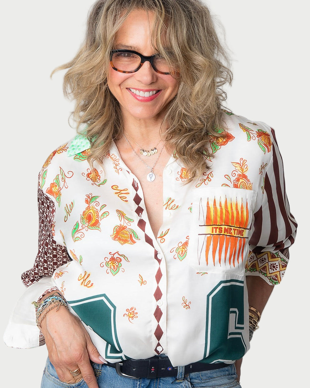 A smiling woman with wavy blonde hair and glasses wears the ME369 Isabel Shirt, a colorful long sleeve button-down with "It's Me Time" on one pocket, paired with layered necklaces, bracelets, and blue jeans against a plain background.