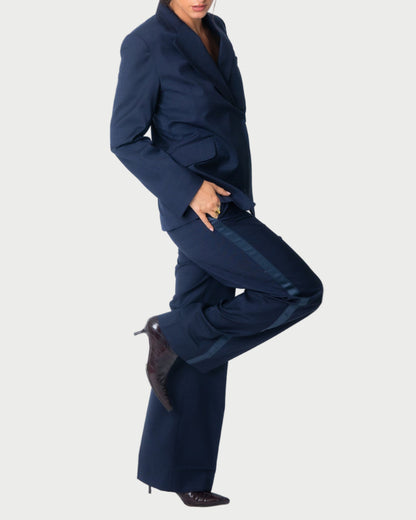 A person in WE-AR4 Tuxedo Trouser and matching blazer stands on one leg, slightly bending the other knee. They wear pointed dark heeled boots against a plain light gray background.